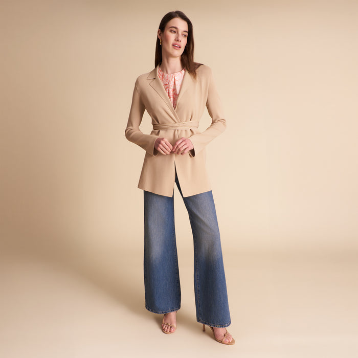 Woman wearing a beige blazer over a light blue shirt and jeans on a plain background | Lead