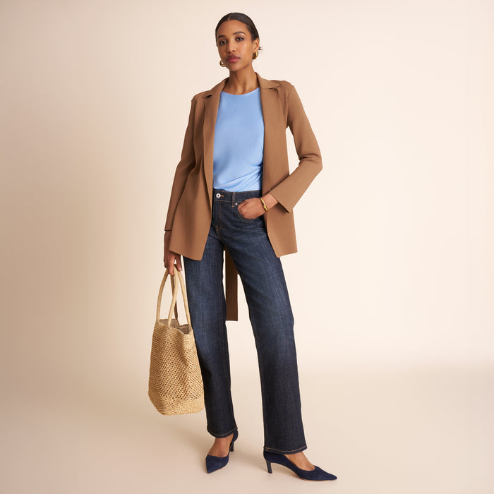 Woman wearing a brown coat, blue jeans, and holding a beige bag on a beige background