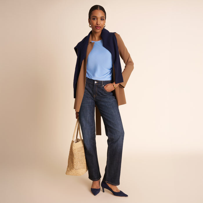 Woman wearing a blue top, dark jeans, and a tan coat holding a woven bag on a beige background