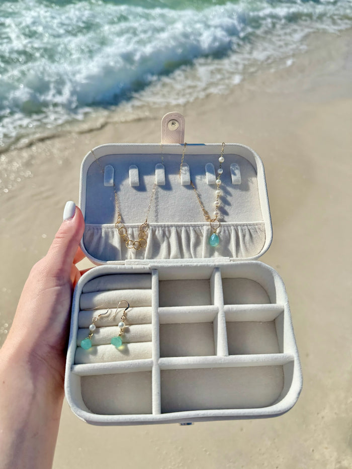 A travel jewelry organizer in a creamy white color, with multiple compartments containing various pieces of jewelry, held open by a hand against a beach and ocean backdrop.