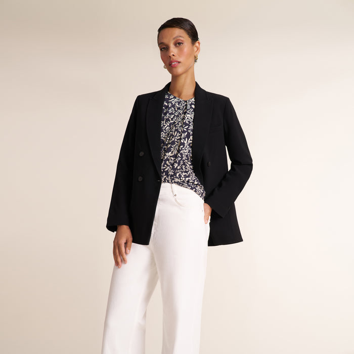 A woman modeling a black double-breasted blazer over a black and white floral print top, paired with white trousers, against a solid light beige studio background.
