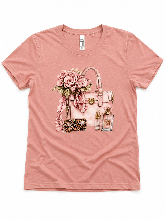 Soft pink short sleeve graphic tee featuring a handbag, floral bouquet, and glam accessory illustration.