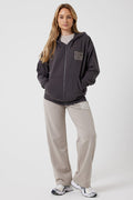 A woman stands against a plain background in the Ralph Street Wide Leg Tracksuit – Grey/Mocha with BC on the chest and leg, a beige t-shirt, and white sneakers. Her hands are tucked into her hoodie pockets.