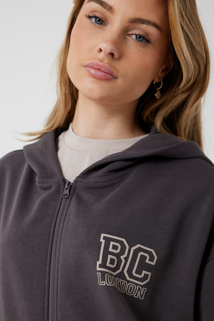 A woman with light brown hair wears the Ralph Street Wide Leg Tracksuit in Grey/Mocha, a dark athleisure full zip hoodie with BC LONDON print, over a beige shirt and gold hoop earrings, looking at the camera neutrally.