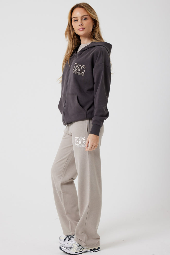 A woman wears the Ralph Street Wide Leg Tracksuit in grey/mocha, featuring a dark gray full zip hoodie and light gray wide-leg joggers with BC text, styled with white sneakers for an effortless athleisure look against a plain light backdrop.