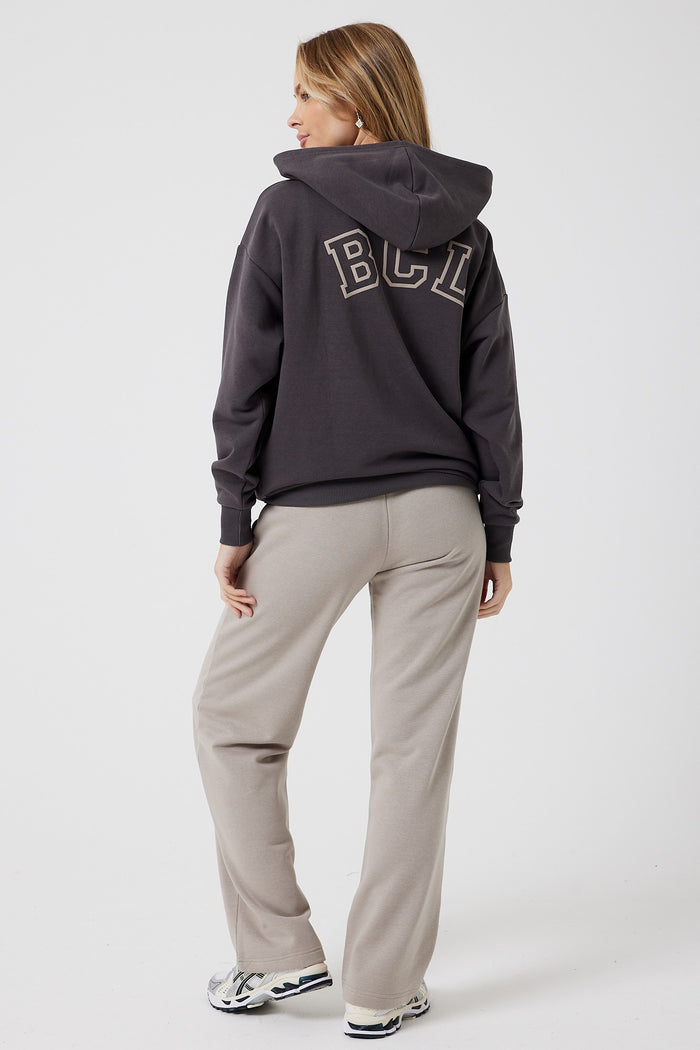 A woman with long, light brown hair faces away, wearing a dark hoodie and the Ralph Street Wide Leg Tracksuit in grey/mocha, paired with white sneakers. She stands against a plain white background.