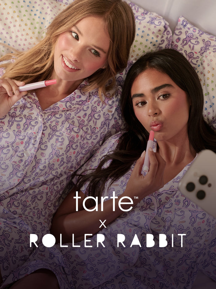 Two women in matching pajamas lying in bed, applying tarte maracuja juicy lip vinyl and looking at a phone. tarte cosmetics x roller rabbit collaboration