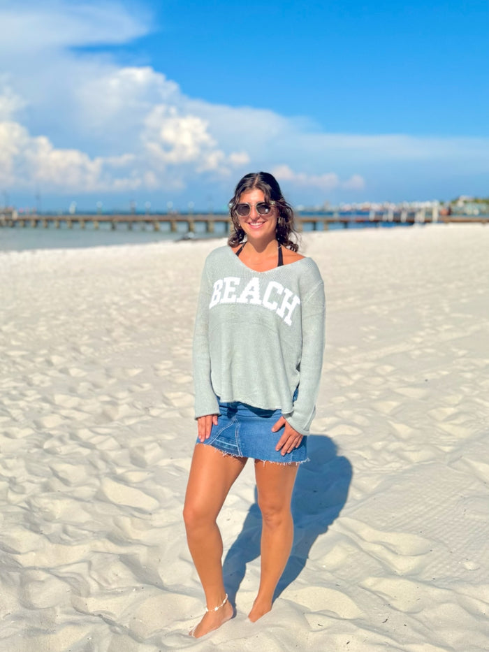 Woman in sunglasses and a 'BEACH' sweatshirt standing on a sandy beach with a clear sky.