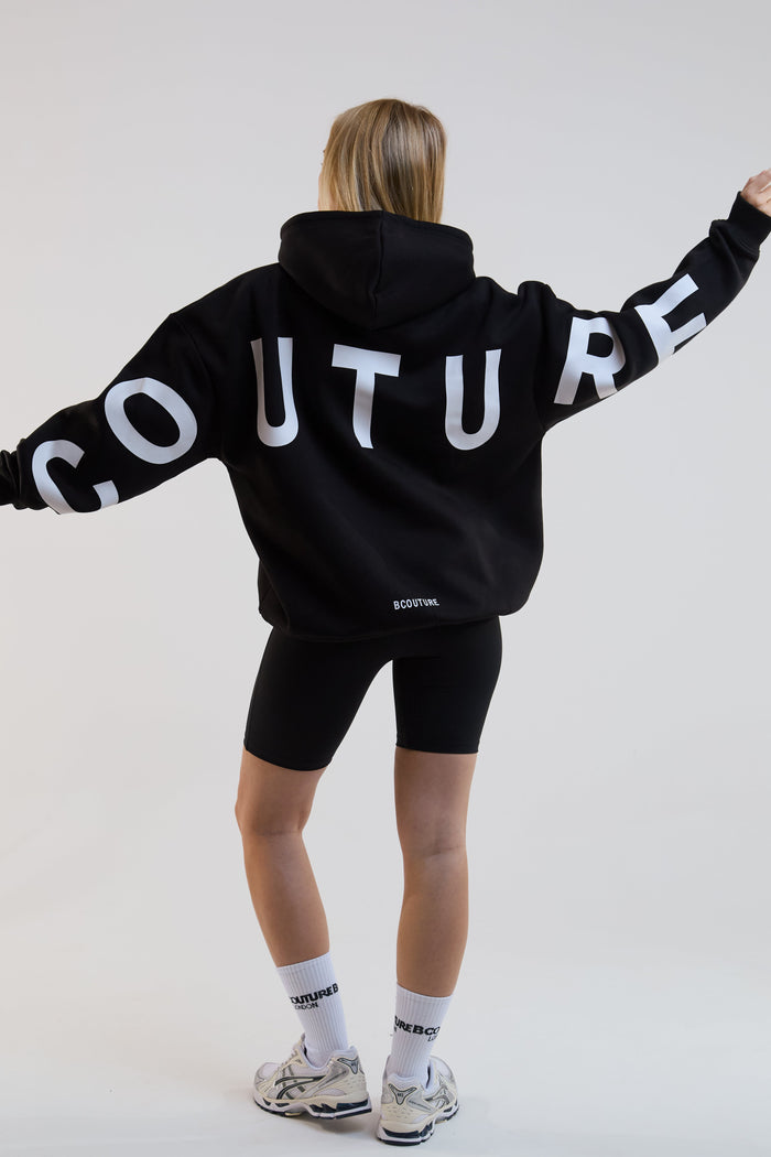 Person wearing a black hoodie with 'COUTURE' text on the back, standing against a white background.
