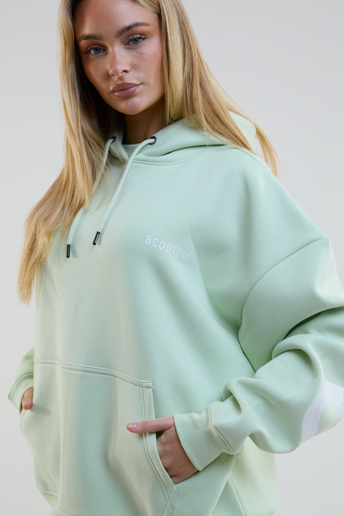 Person wearing a light green hoodie with a brand logo on a plain background