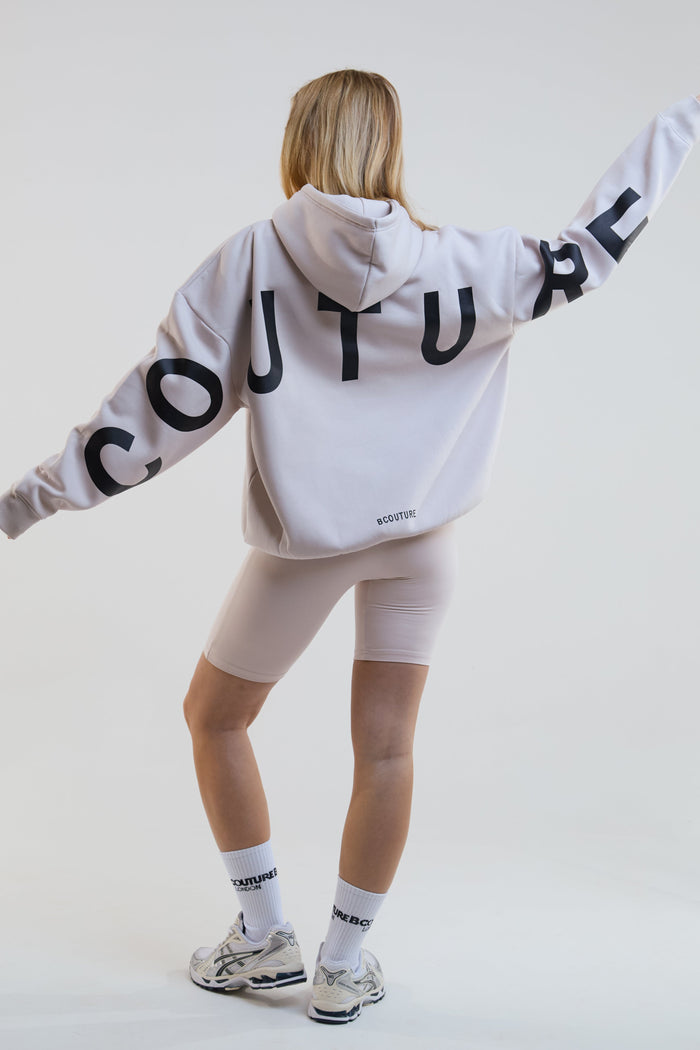 Person wearing a beige hoodie with 'COUTURE' text on the back, standing against a plain background.