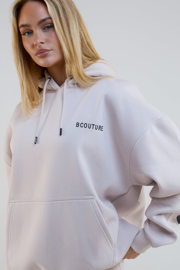 Woman wearing a light beige hoodie with 'BCOUTURE' branding on a white background