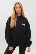 A woman with long blonde hair wears the Westwood Oversized Hoodie - Black, featuring white chest text and a loose fit, paired with black leggings while posing in front of a plain white background.
