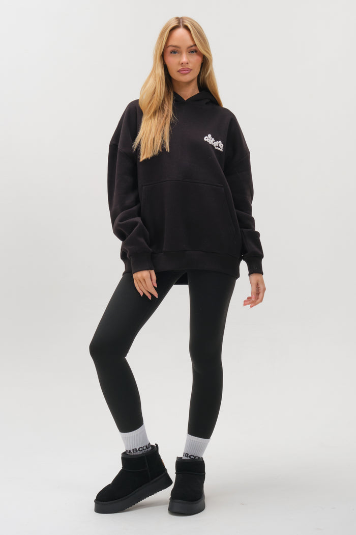 A woman with long blonde hair stands against a plain background, wearing the Westwood Oversized Hoodie in black. She pairs it with black leggings, slip-on shoes, and white socks, looking at the camera with a relaxed expression and hand on her hip.