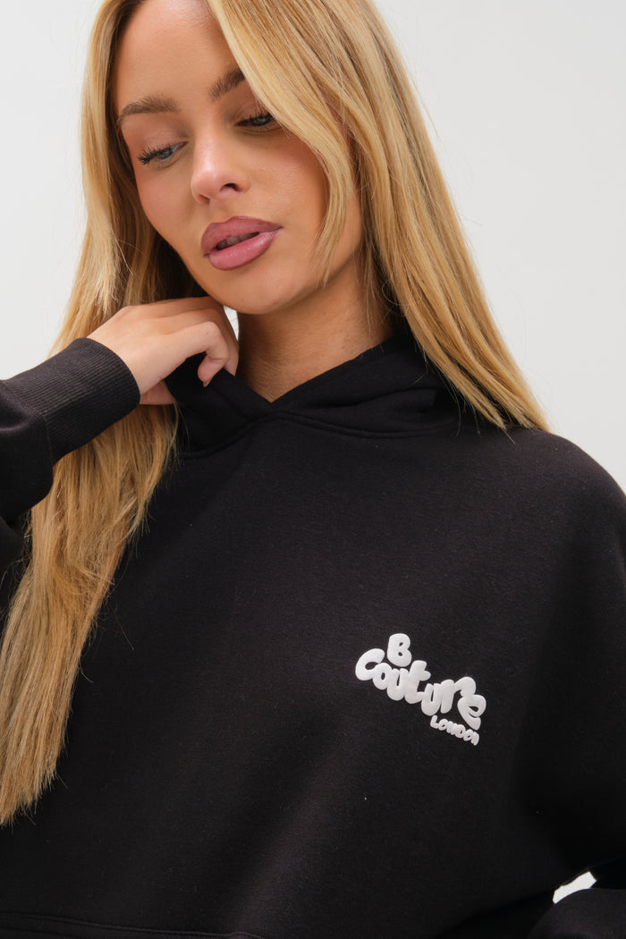 A woman with long blonde hair wears the Westwood Oversized Hoodie - Black, featuring "Be Couture London" in white on the chest. She looks down and touches her hair against a plain light background.