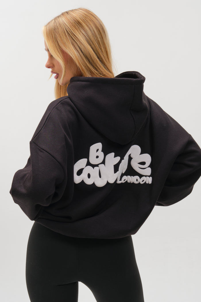 A woman with long blonde hair wears the Westwood Oversized Hoodie - Black, featuring bold white "B COURTIE LONDON" lettering on the back, paired with black leggings. She stands facing away from the camera against a plain background.
