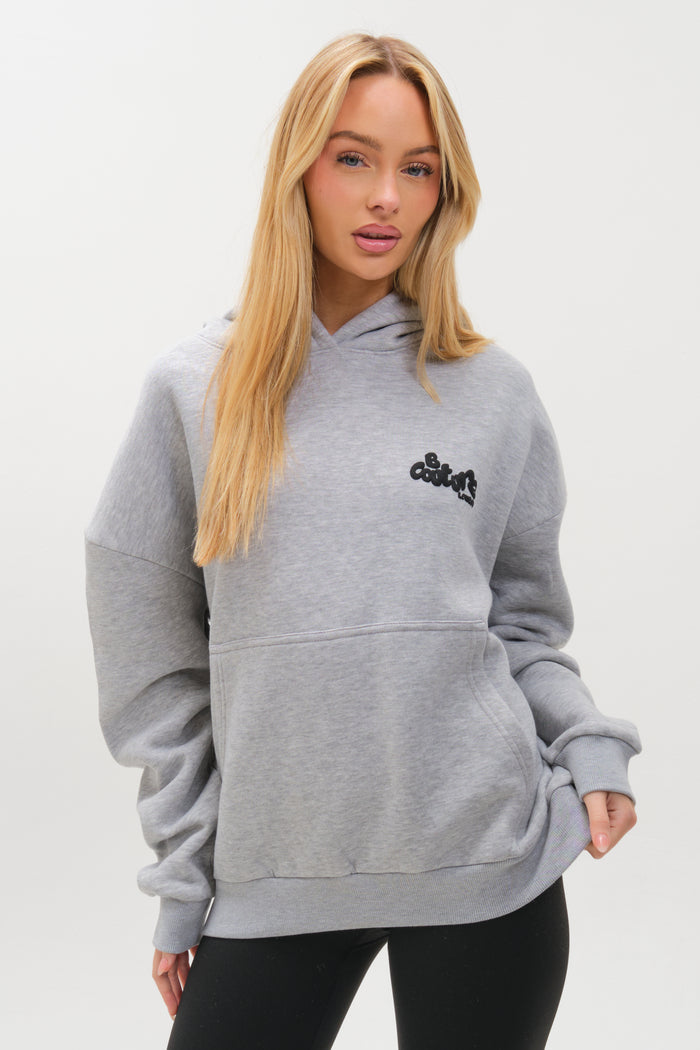A woman with long blonde hair models the Westwood Oversized Hoodie in Grey Marl, featuring black chest lettering and a comfy fit, paired with black leggings against a plain white background.