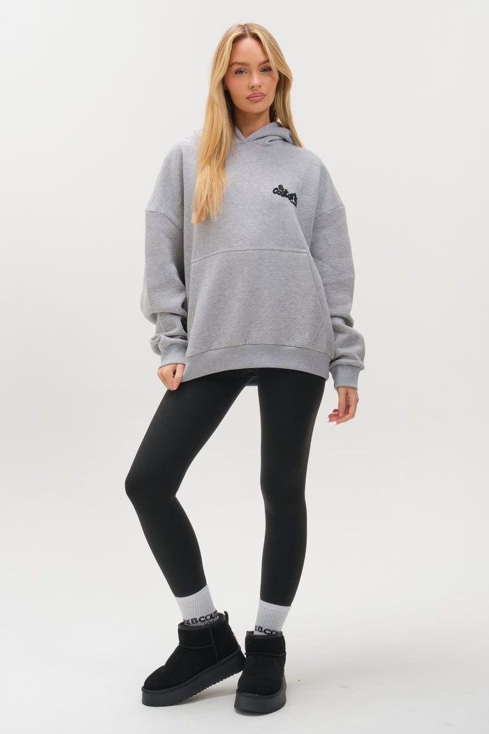 A woman with long blonde hair models the Westwood Oversized Hoodie - Grey Marl. She stands against a white background, wearing black leggings and black shoes, showing off the hoodie’s relaxed fit and comfort.