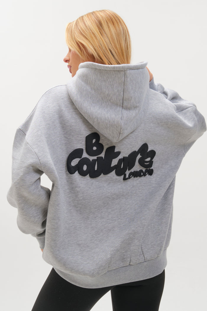 A person with long blonde hair wears the Westwood Oversized Hoodie - Grey Marl, its hood up and bold black "B Couture London" text on the back. The relaxed, cozy fit is showcased as they face away from the camera.