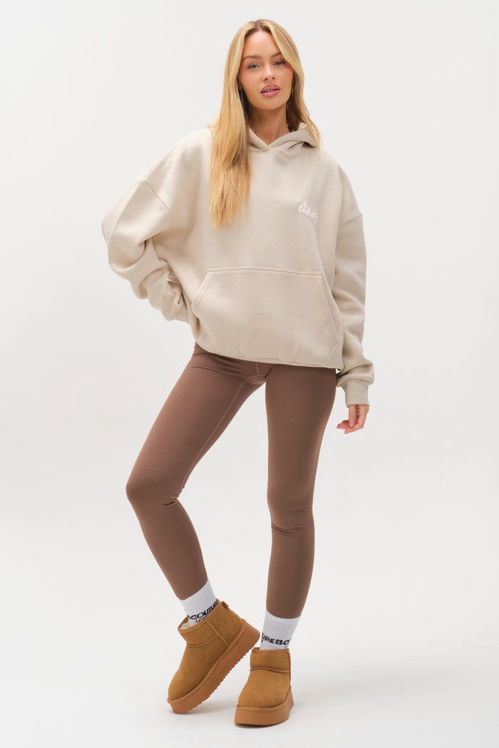 A woman poses against a white background wearing the Westwood Oversized Hoodie - Stone, paired with brown leggings, white socks, and tan ankle boots. She has long blonde hair and stands with one leg slightly bent and a hand in her hoodie pocket.