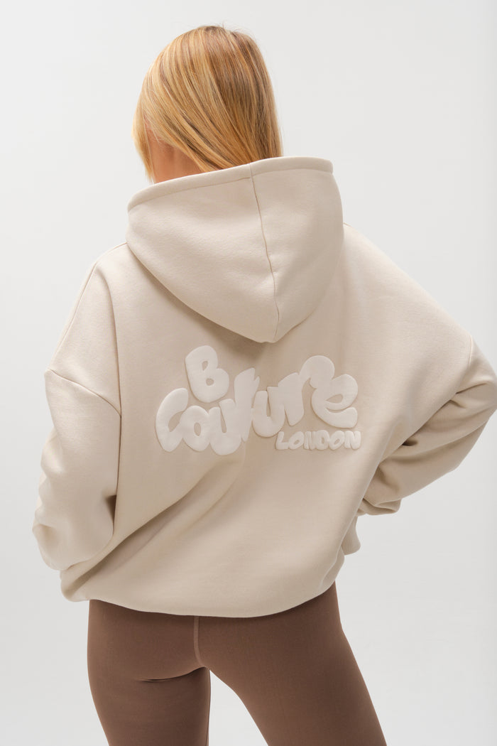 A person with long, light brown hair is seen from behind wearing the Westwood Oversized Hoodie - Stone, a relaxed fit beige cotton blend piece featuring "B Couture London" in white on the back, paired with brown leggings against a white background.