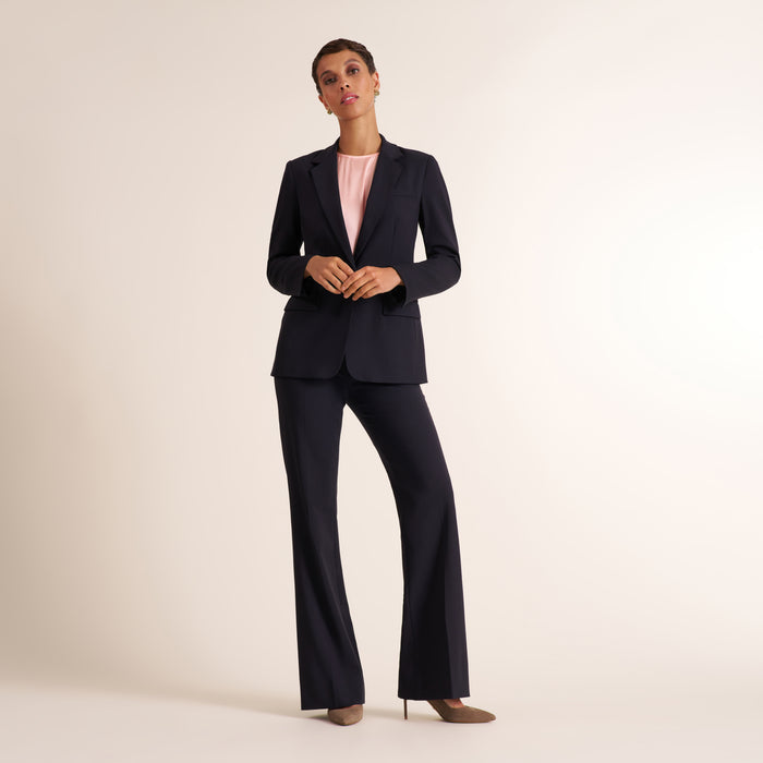 A model stands in a studio wearing a dark navy blue two-piece suit consisting of a blazer and matching wide-leg trousers, layered over a light pink top and styled with brown pointed-toe heels. | Lead