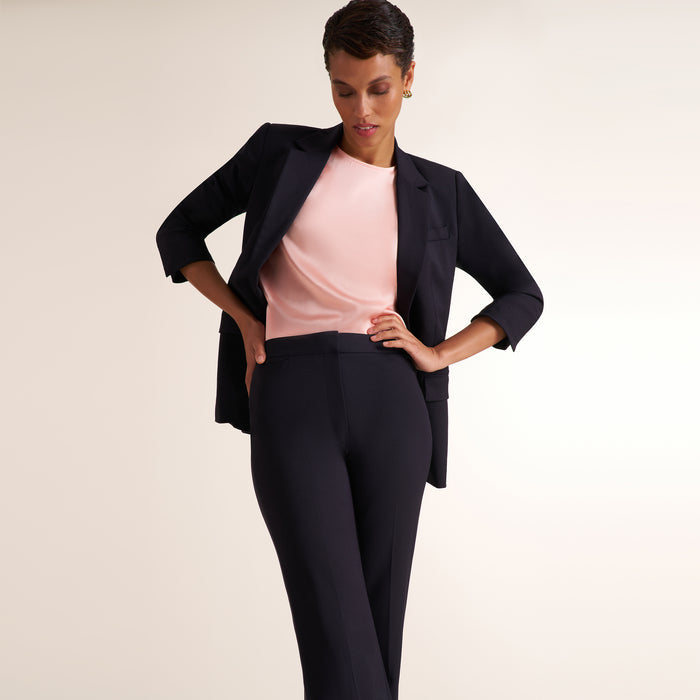 A woman modeling a dark navy blue tailored suit consisting of a blazer and trousers, worn over a light pink top, against a plain light beige studio background.
