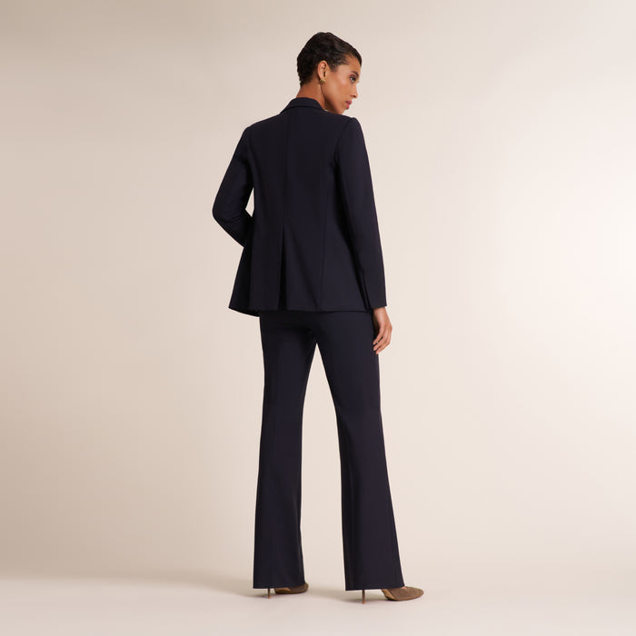 A model is shown from the back wearing a dark navy blue tailored suit, consisting of a blazer and matching wide-leg trousers, against a solid light beige studio background.