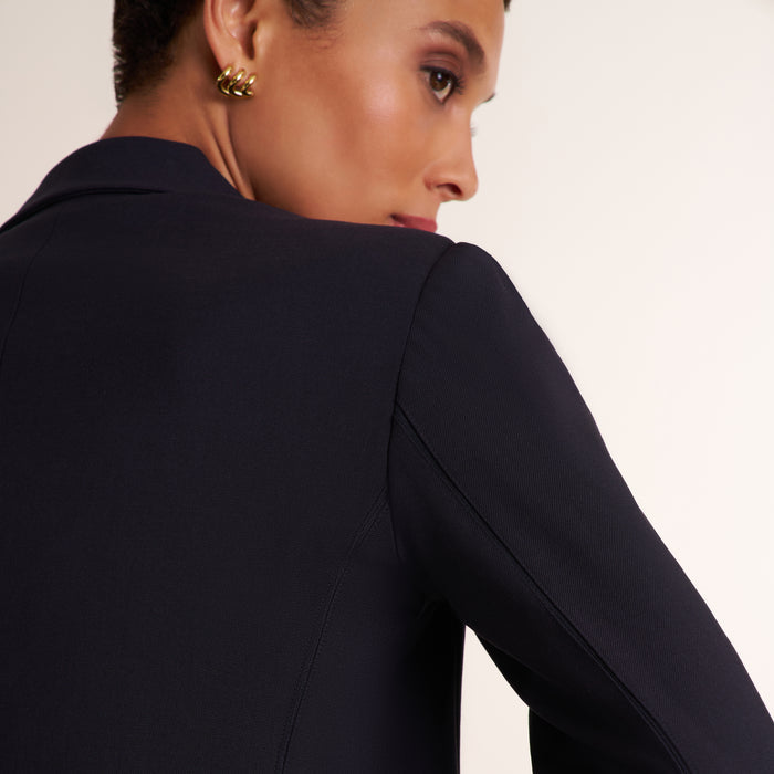 A close-up side profile of a model wearing a dark navy blue blazer and a textured gold earring against a plain off-white background.